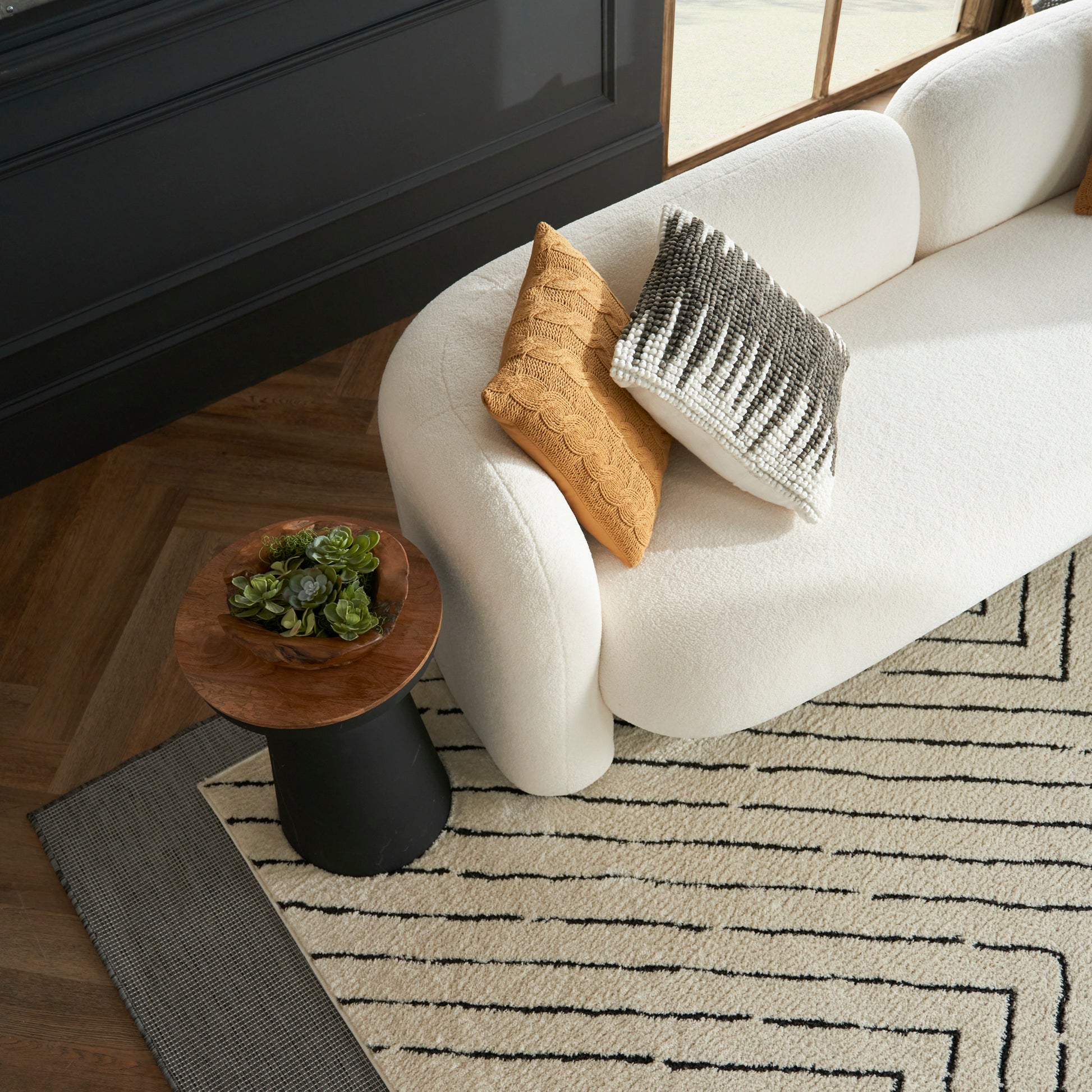 Cream curved sofa with mustard knit and black-to-white gradient pillow, round wood side table with succulents, geometric rug.