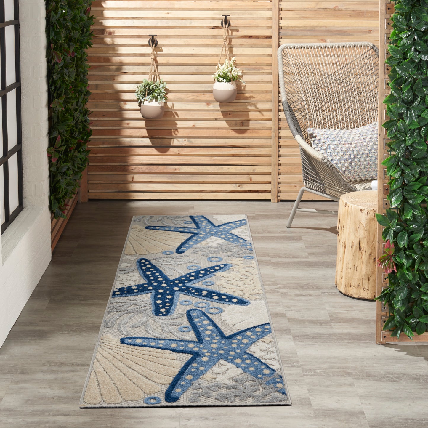 Outdoor runner rug featuring blue starfish pattern on beige background, laid on wooden deck beside wicker chair and planters.