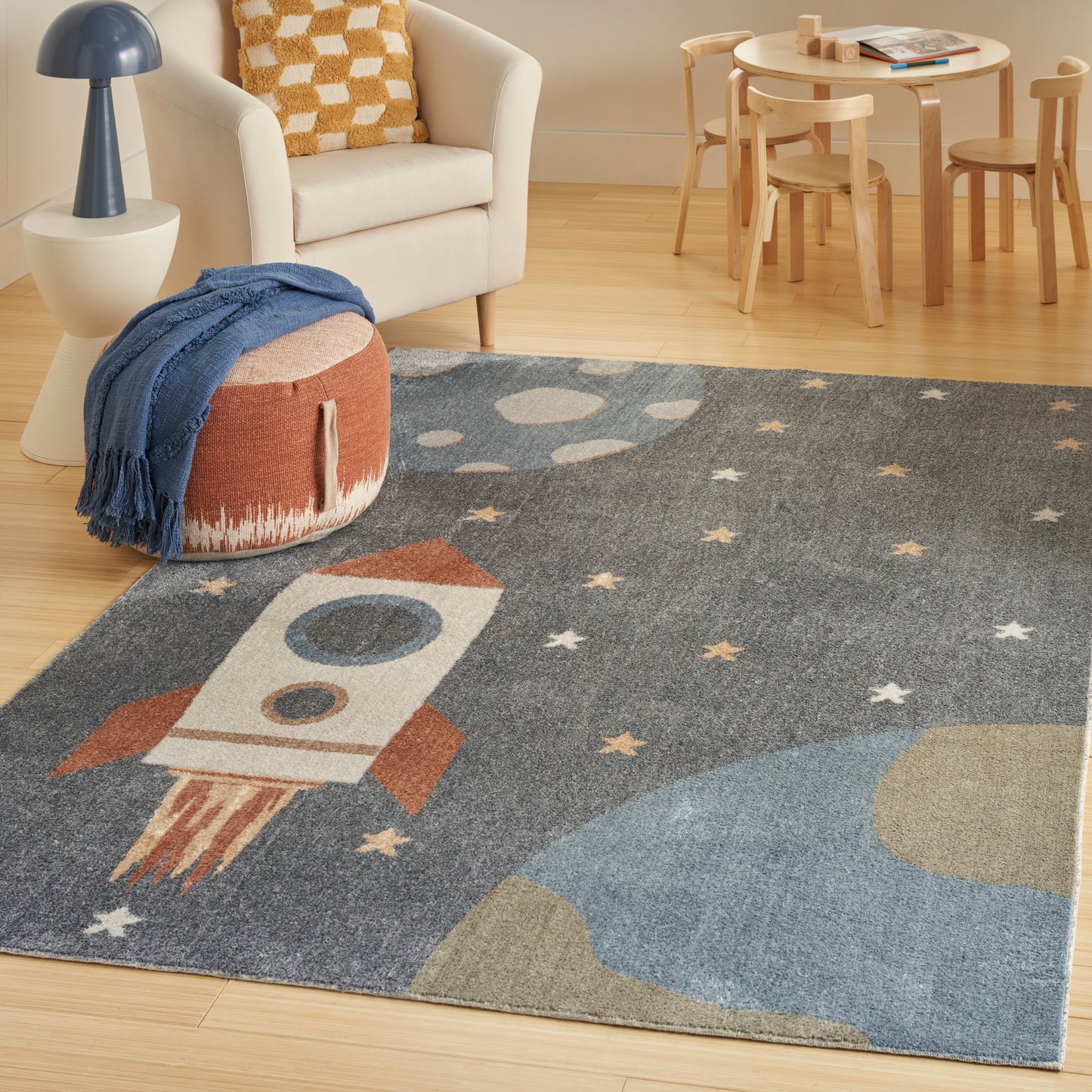 Soft blue-gray kid's rug featuring a white-and-orange rocket, stars and planets; cozy play area with a white chair and wooden table.