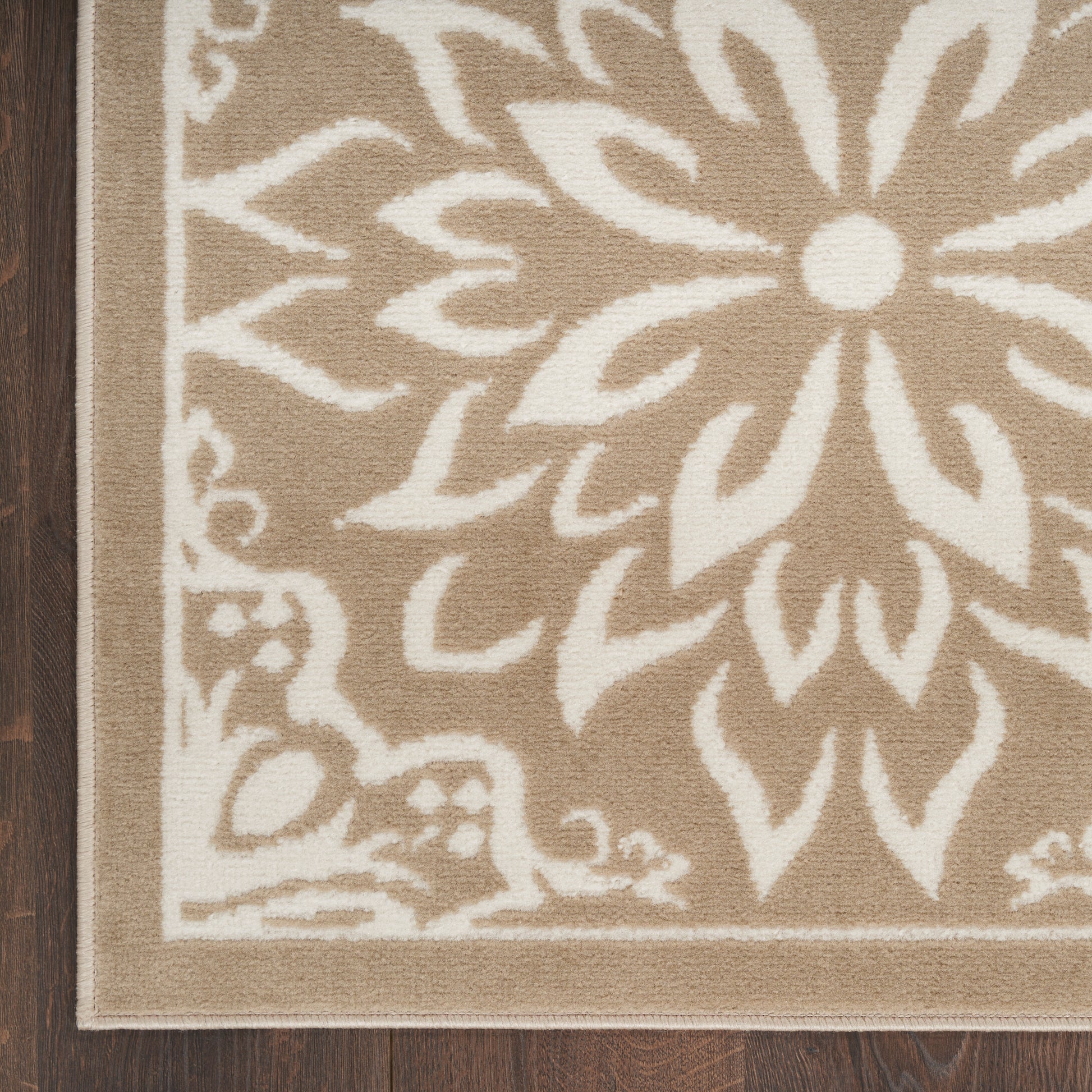 Beige woven area rug with large white floral motif & border, on dark wood floor.