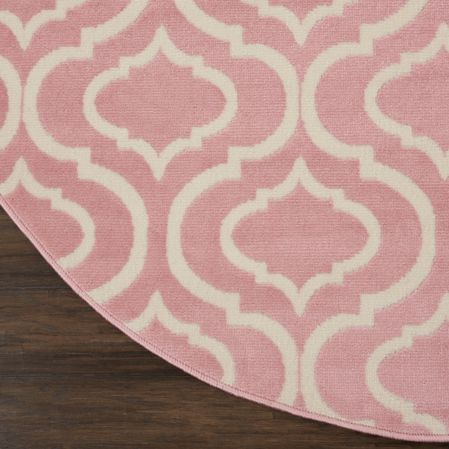 Pink rug with white Moroccan-inspired trellis pattern, curved edge on dark wooden floor.