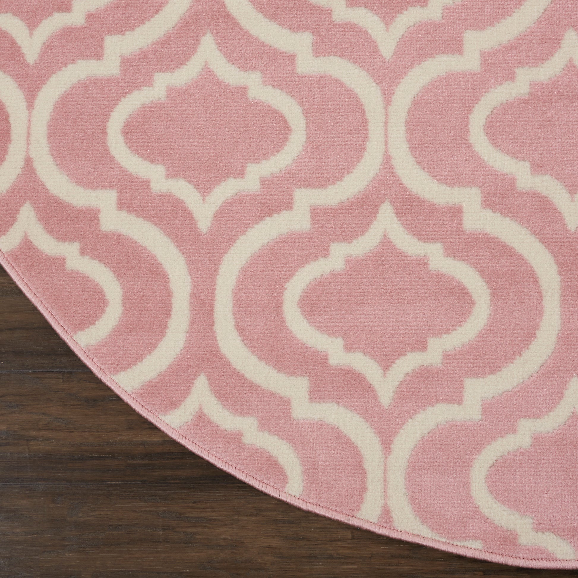 Pink rug with white Moroccan-inspired trellis pattern, curved edge on dark wooden floor.
