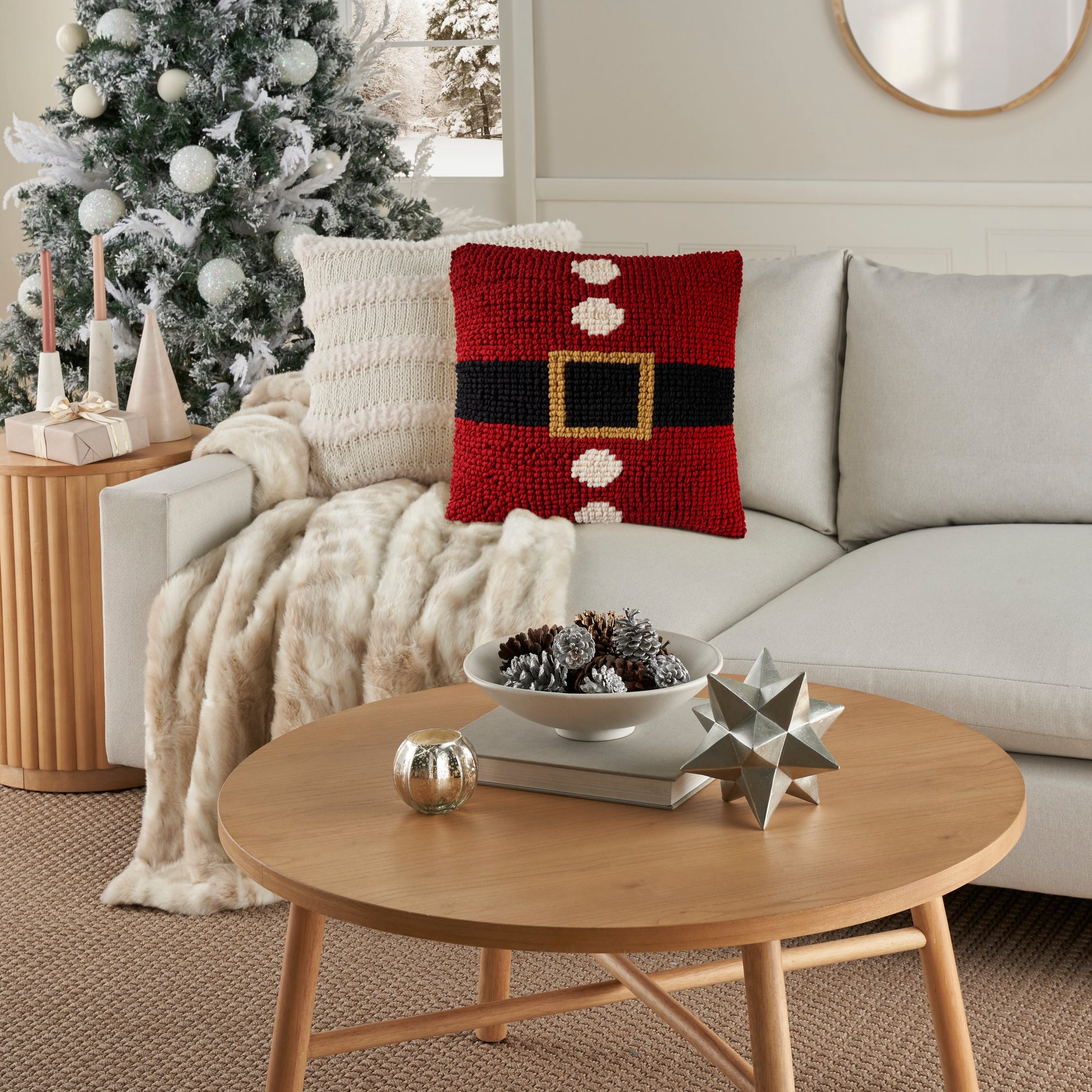 Cozy living room with Santa belt pillow on light sofa, cream throw, frosted tree, round wooden coffee table with metal stars