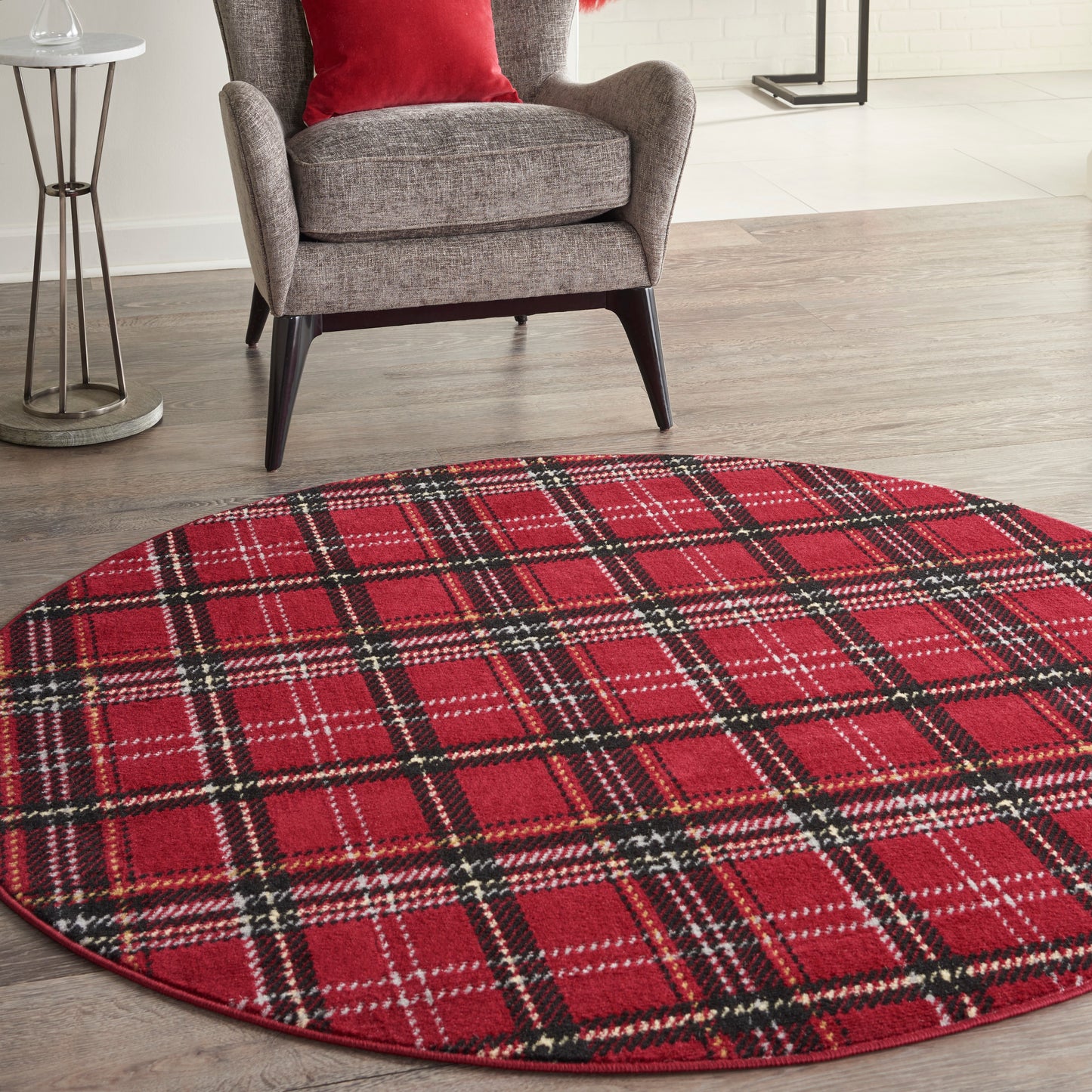 Round red plaid rug on dark wood floor beside a gray upholstered armchair with a red pillow.