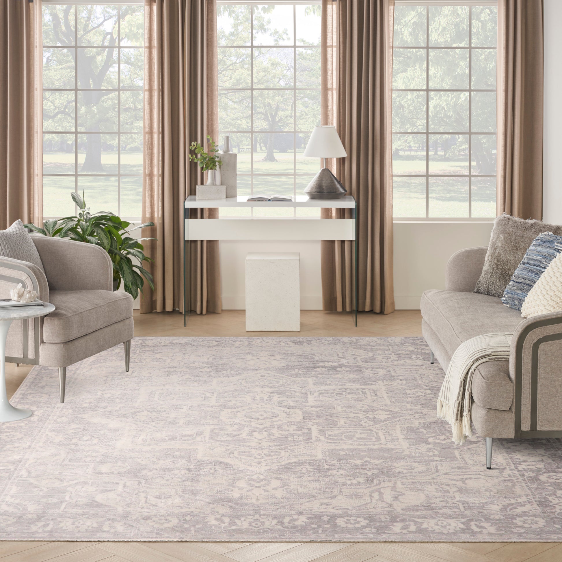 Neutral living room with beige sofas, taupe curtains, glass-top console, gray patterned rug and potted greenery.