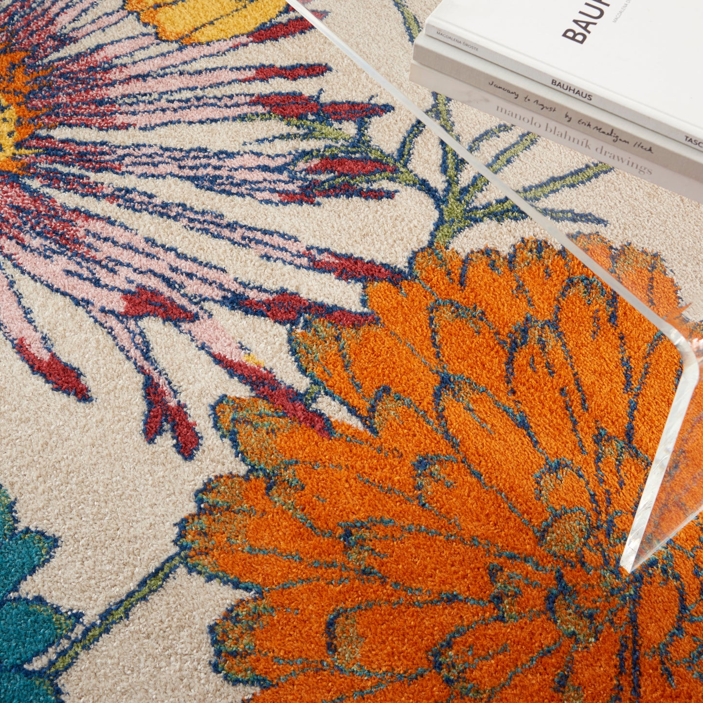 Vibrant floral rug with a large orange bloom, purple stems, blue-green accents; acrylic coffee table edge and stacked books.
