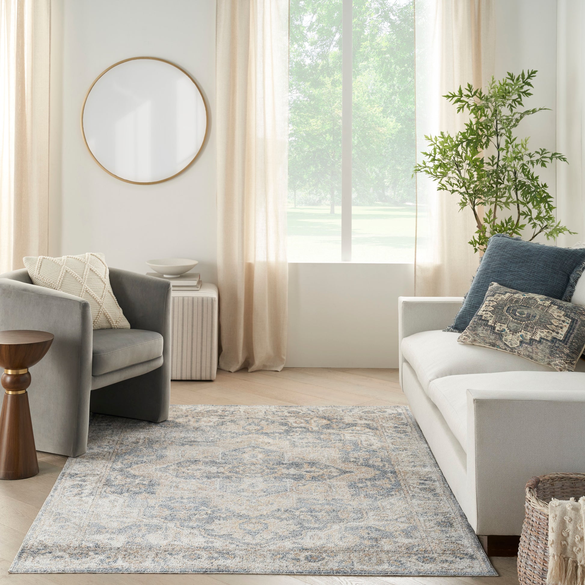 Bright living room with round wooden-framed mirror, beige curtains, blue-gray patterned rug, gray sofa and chair, lush plant.