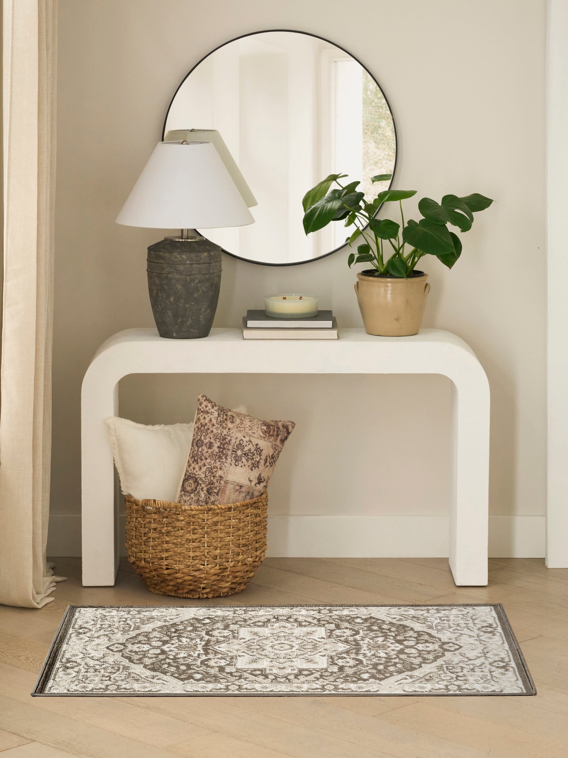 Round mirror above a white curved console with a stone lamp, potted plant, books and a woven basket on a gray patterned rug.
