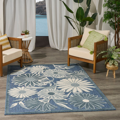 Blue floral area rug with white daisies in tropical living room, wooden-framed chairs, beige cushions, and green plants.