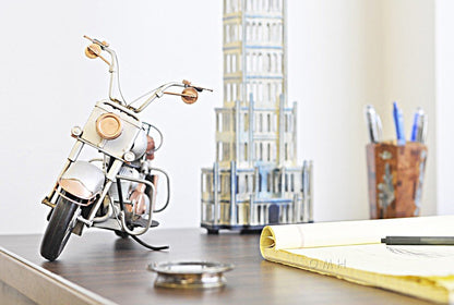 Miniature metal motorcycle desk ornament in silver and copper tones, with a tall tower model and rolled blueprints nearby.