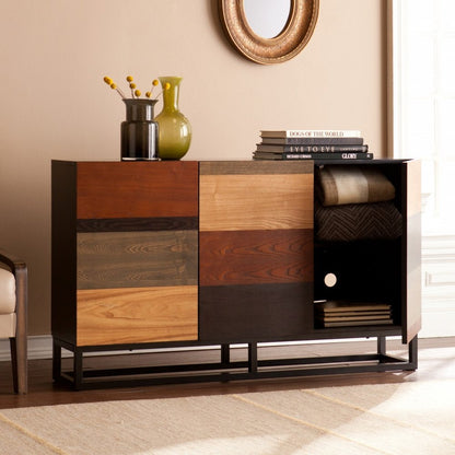 Mid-century wood sideboard with brown gray and black patchwork panels, black frame; top with vases and books, three-door cabinet.