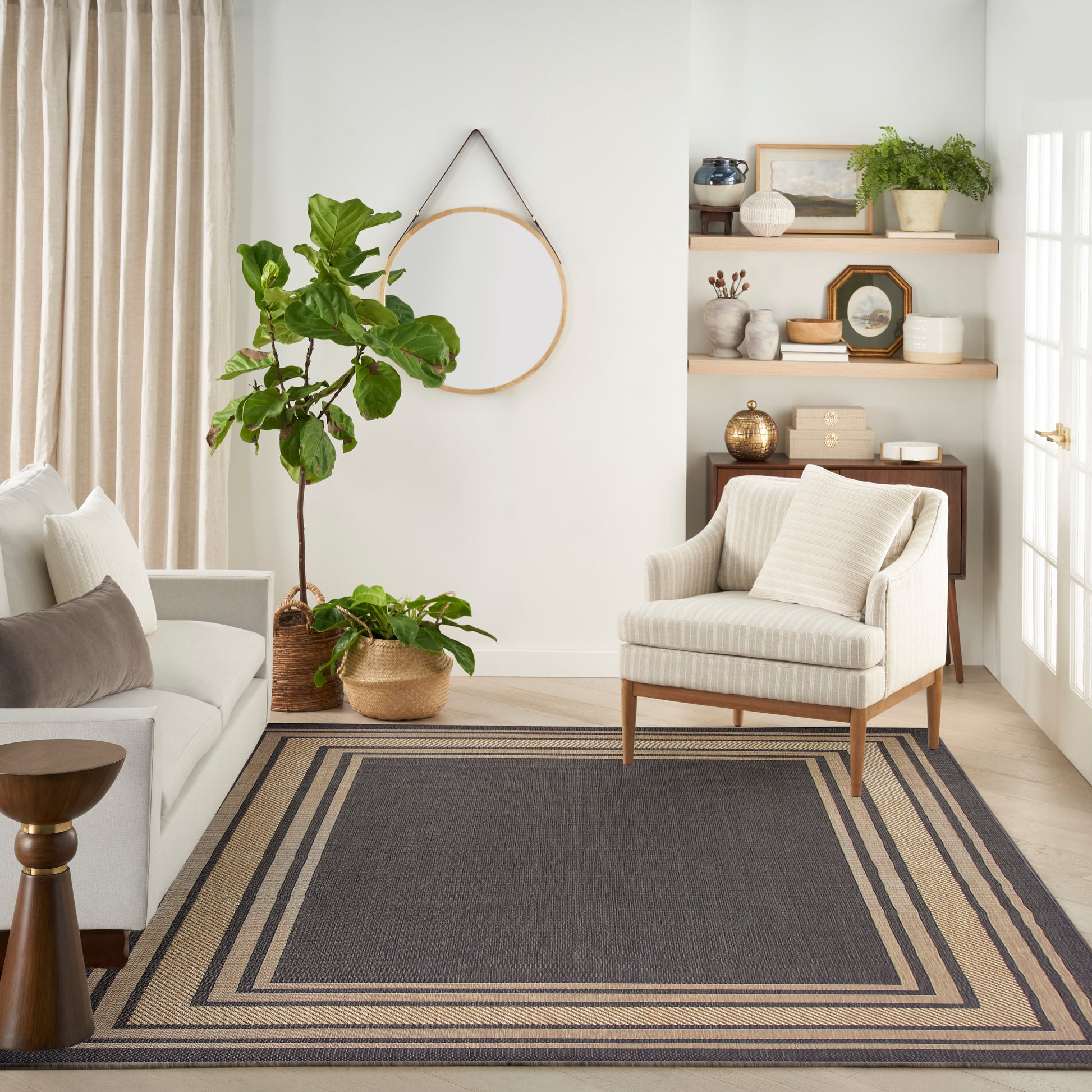 Neutral living room with beige striped armchair, gray sofa, rug, fiddle-leaf fig plants, round wall mirror, wooden shelves.