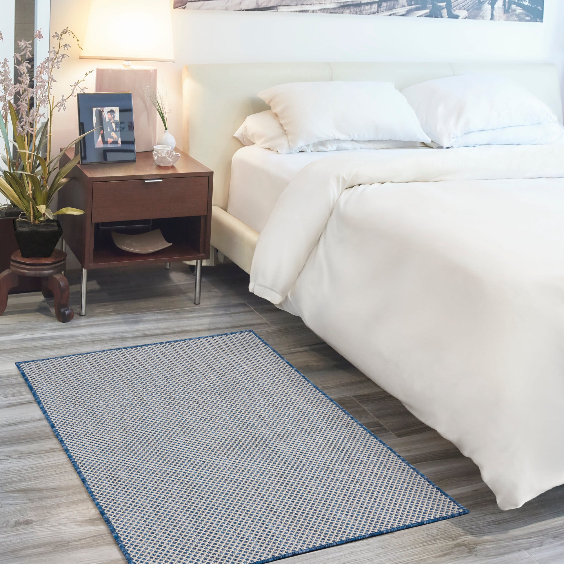 White bedding on a bed with white pillows, dark wood nightstand, lamp, plant, and a blue-bordered woven rug.