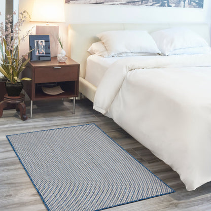 White bedding on a bed with white pillows, dark wood nightstand, lamp, plant, and a blue-bordered woven rug.