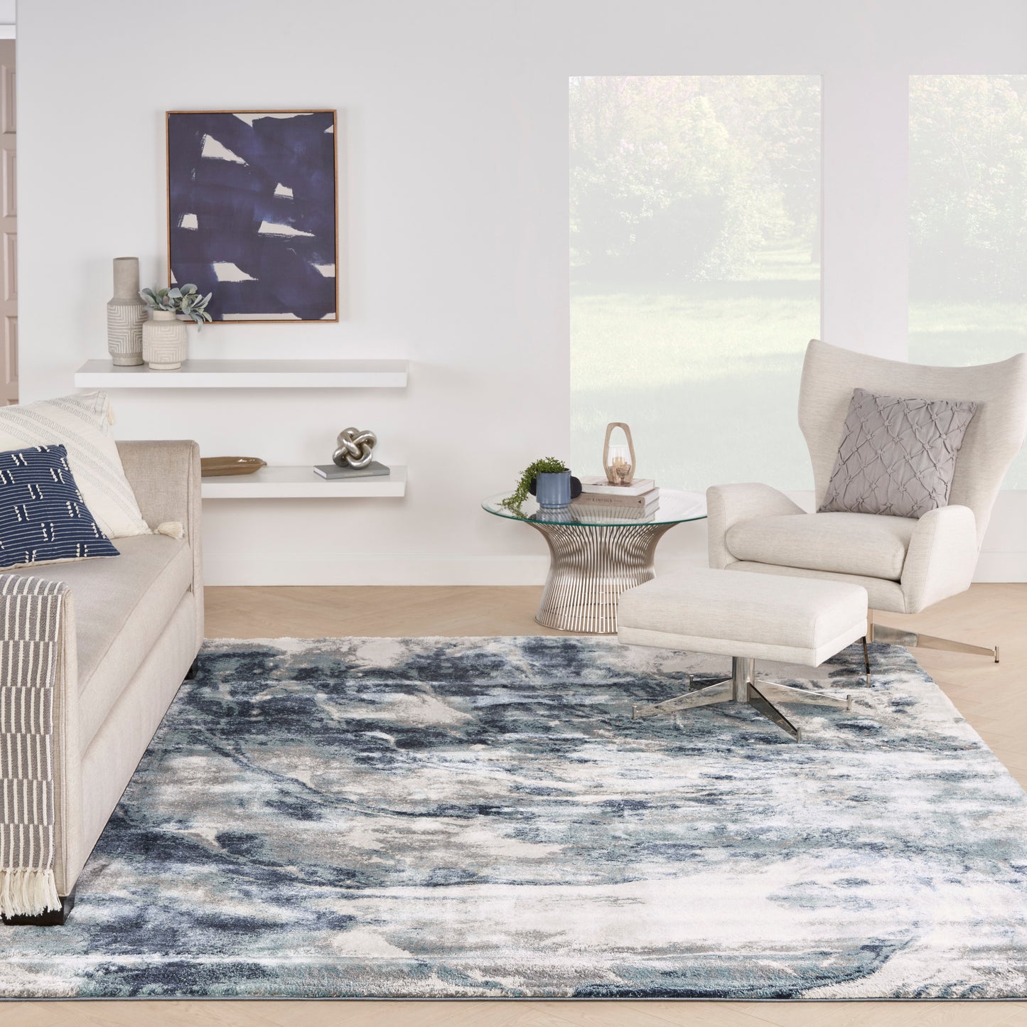 Bright living room with a blue abstract rug, cream sofa and armchair with ottoman, round glass coffee table, and coastal decor.