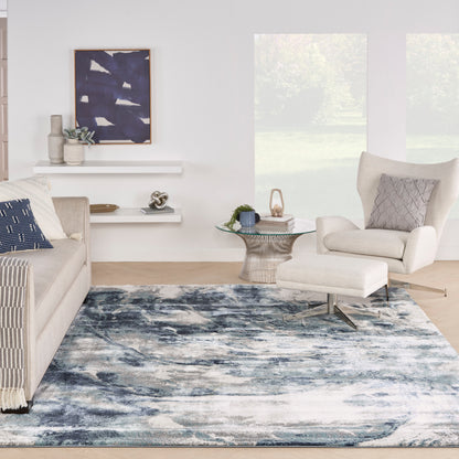 Bright living room with a blue abstract rug, cream sofa and armchair with ottoman, round glass coffee table, and coastal decor.