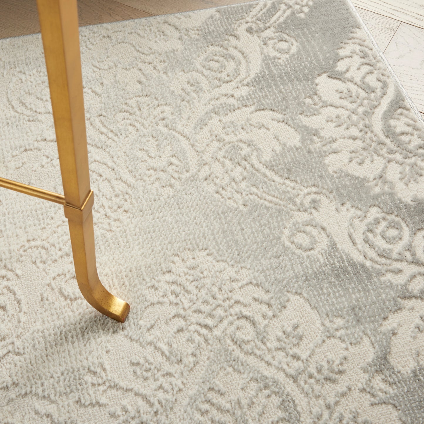 Ivory-gray damask rug with floral scroll pattern; gold metal table leg resting on it.