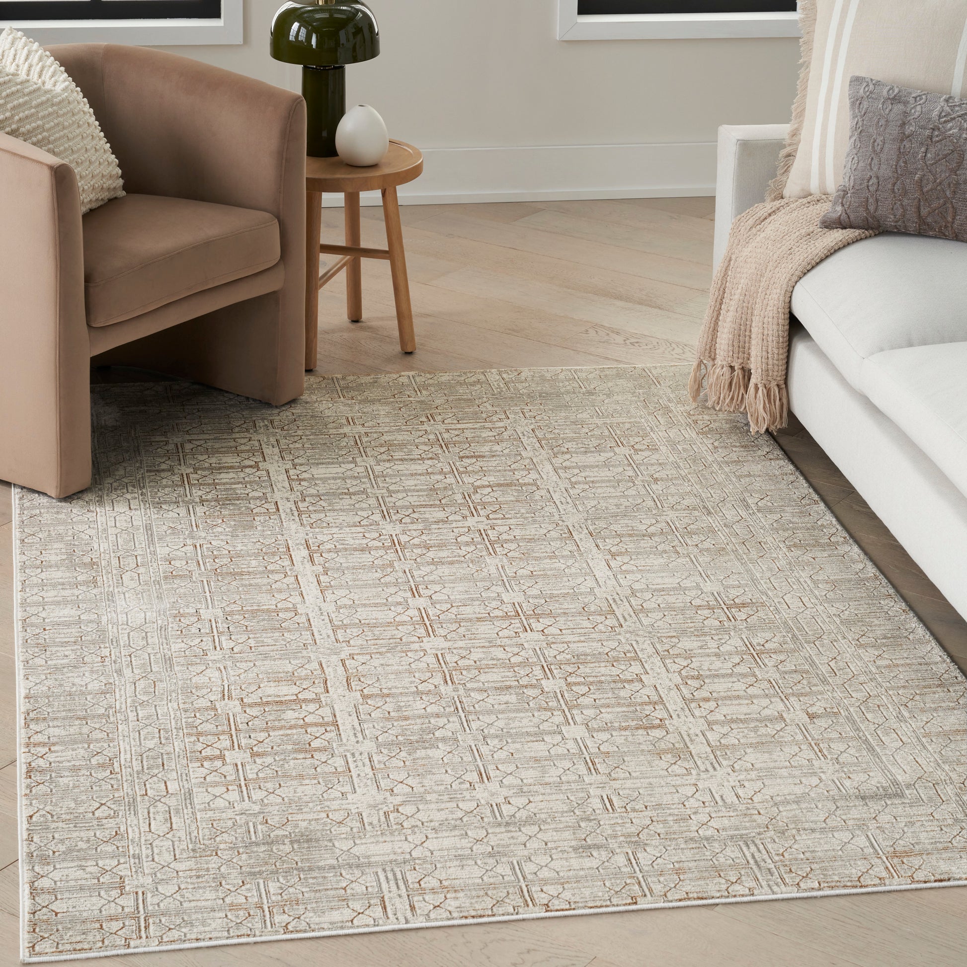 Beige geometric woven area rug on light wood floor with taupe armchairs, white sofa, and round wooden side table.