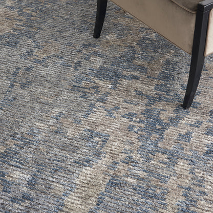 Blue and beige distressed-pattern area rug with abstract motif; partial view of a tan-upholstered chair with black legs.