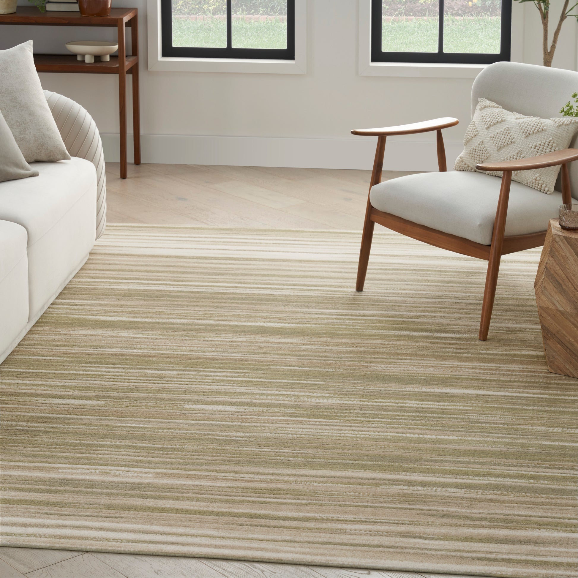 Living room with a beige striped area rug, light sofa, mid-century wooden armchair & round side table by bright windows.
