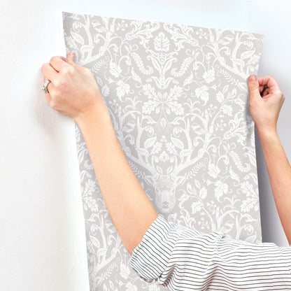 Hands hold a gray damask wallpaper sample against a pale wall; striped shirt sleeves visible.
