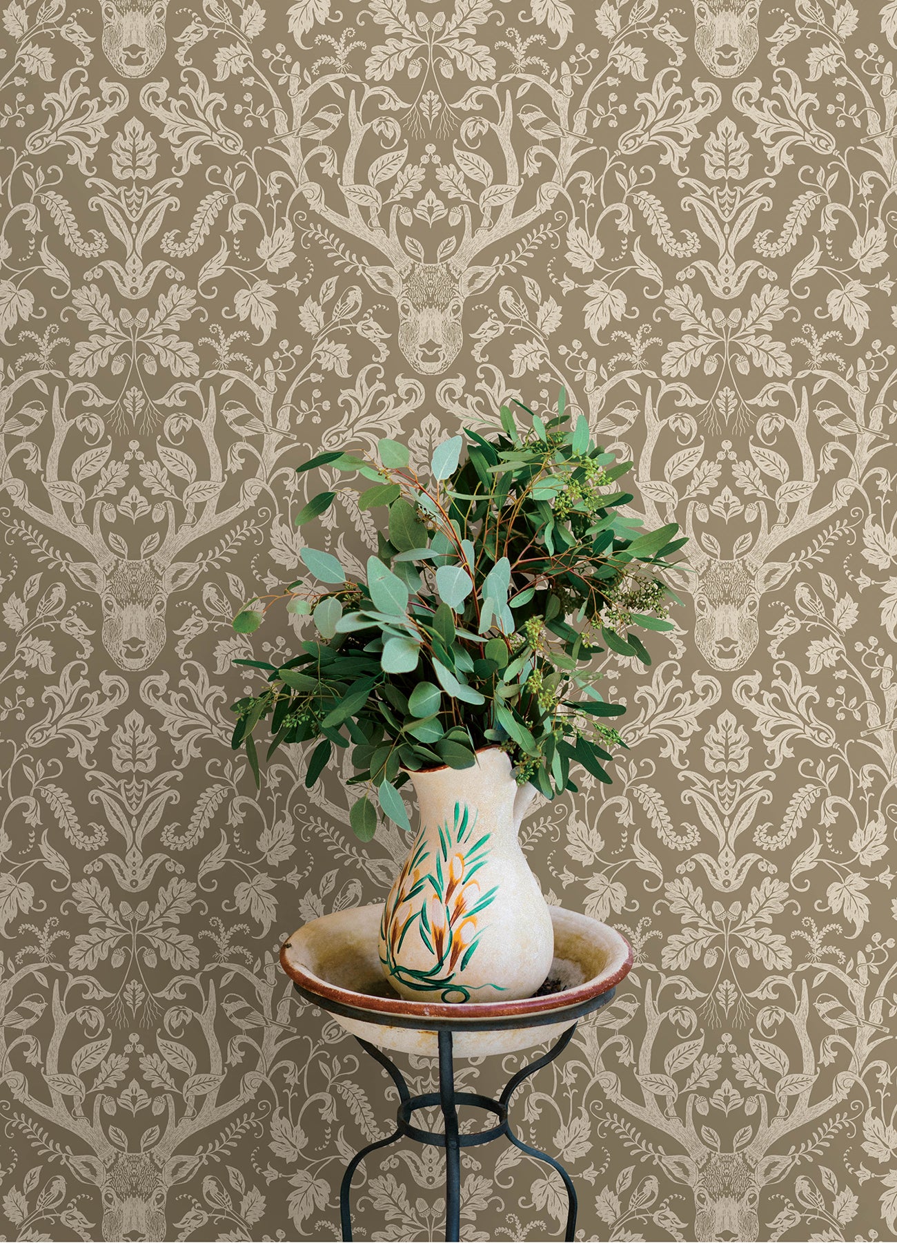 Beige damask wallpaper with deer motif; cream vase with teal and orange leaf design holding eucalyptus on a wrought-iron stand.