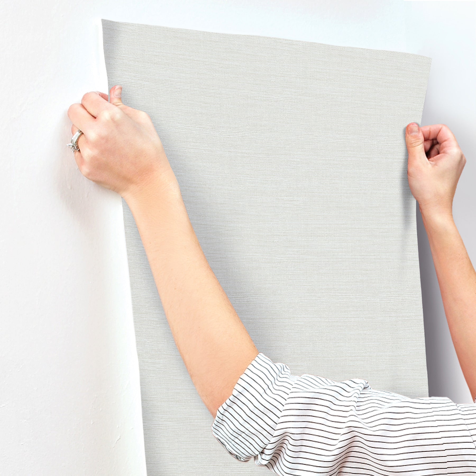Two hands stretch a light blue faux grasscloth wallpaper sample against a wall.