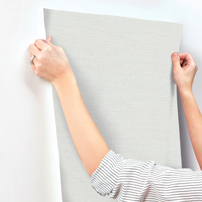 Two hands stretch a light blue faux grasscloth wallpaper sample against a wall.