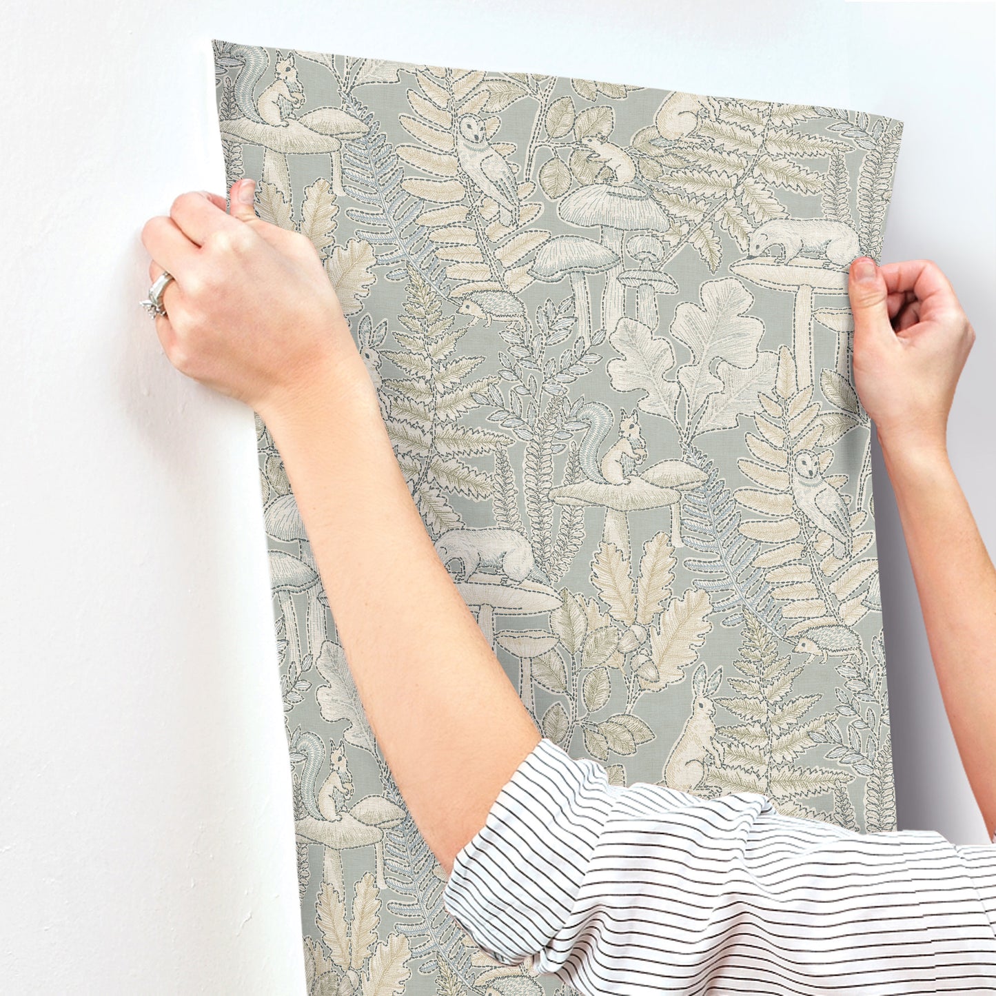 Hands hold up gray wallpaper panel with cream botanical fern pattern; striped shirt cuffs visible.