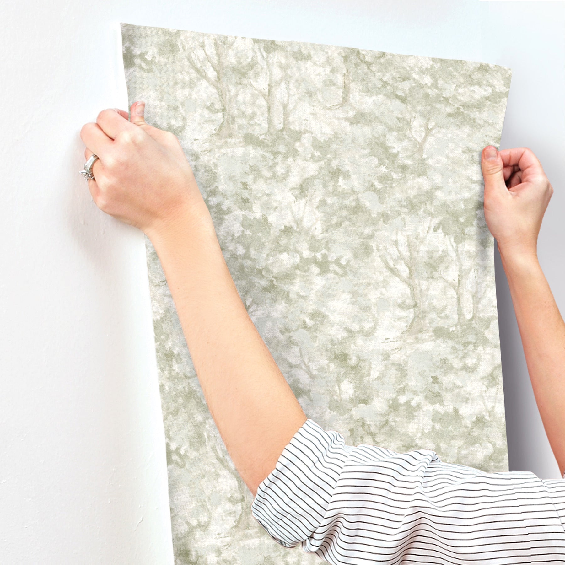 Hands in a striped shirt hold a large gray-green forest/tree wallpaper panel for wall decor.