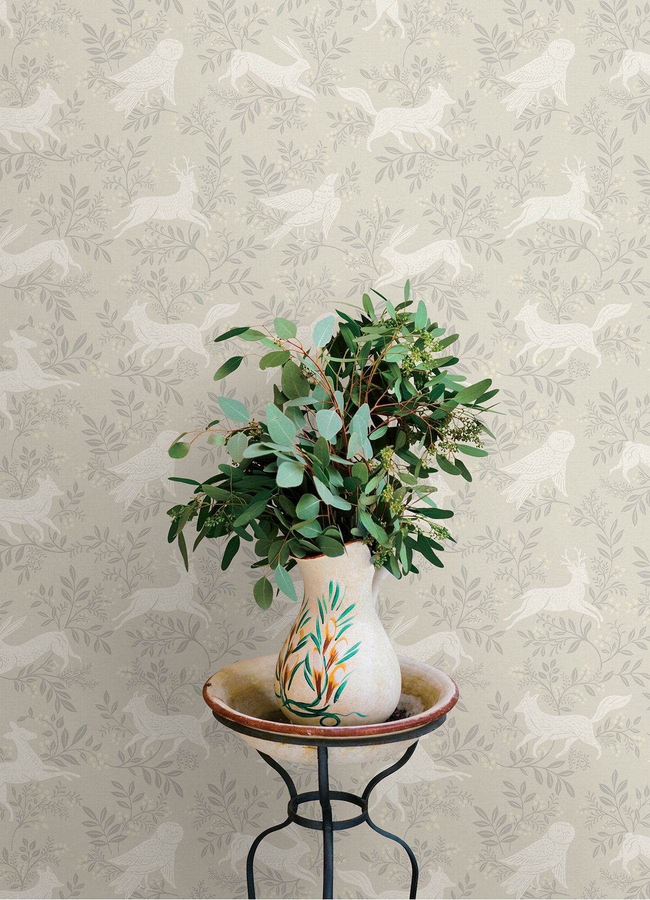 Beige ceramic vase with a botanical motif on a small stand, filled with eucalyptus against pale gray bird wallpaper.