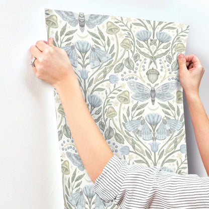 Hands hold a botanical wallpaper panel with blue-green leaves and moth motifs on a white background.