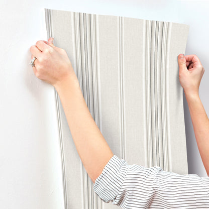 Beige wallpaper sample with vertical gray stripes; embossed texture, held by hands for interior wall decor.
