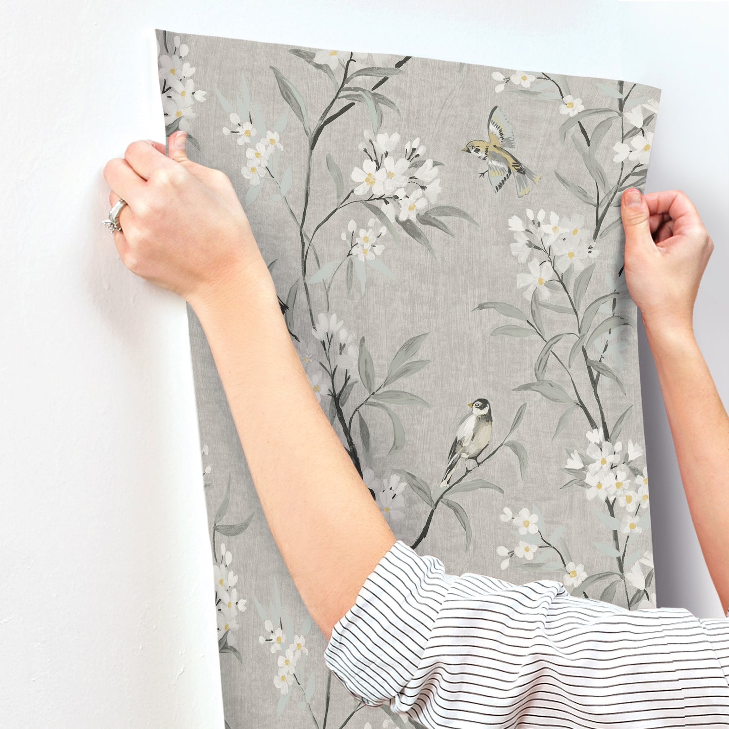 Gray botanical wallpaper panel featuring white blossoms and small birds, held by hands.