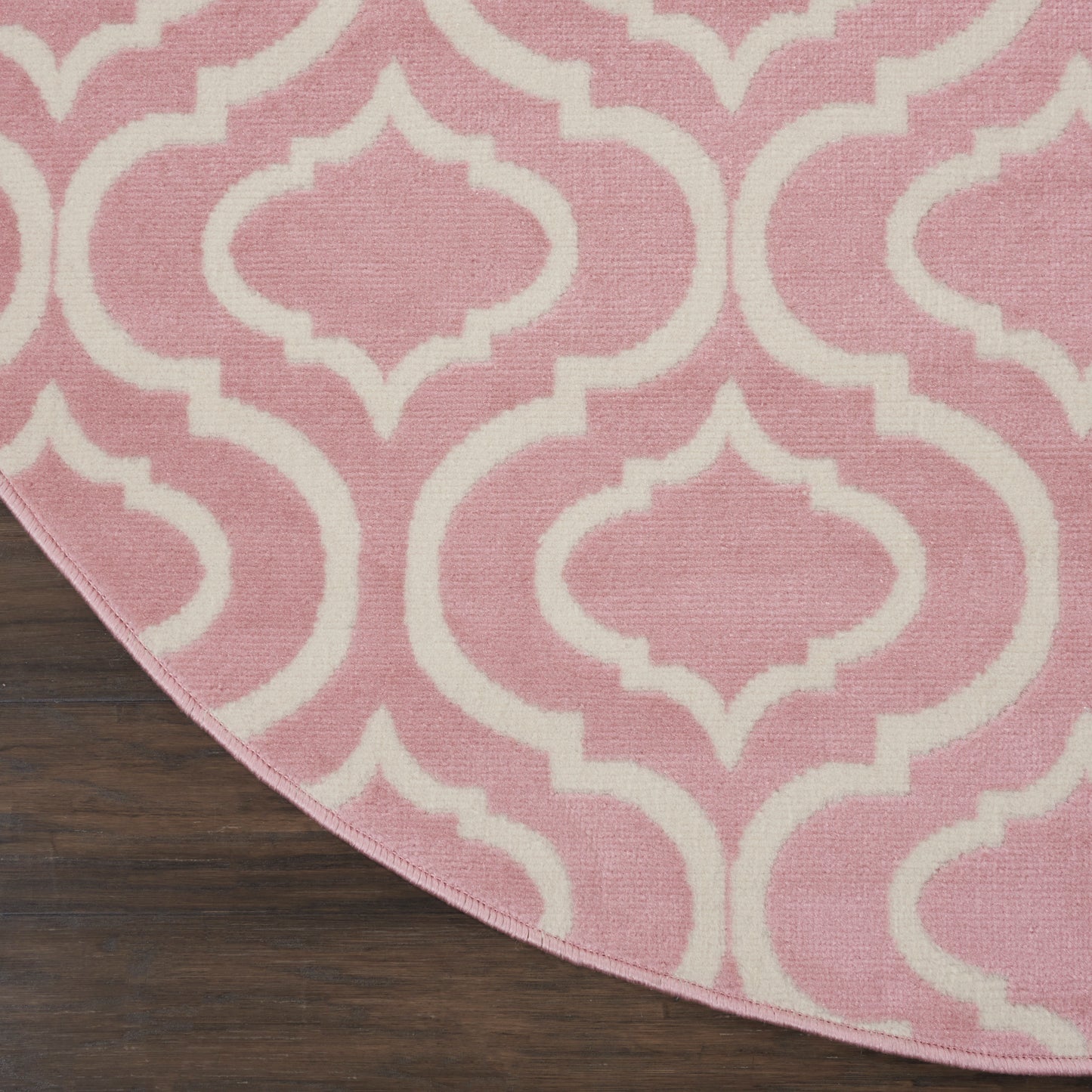 Pink round rug with white trellis pattern; woven texture on dark wood floor.
