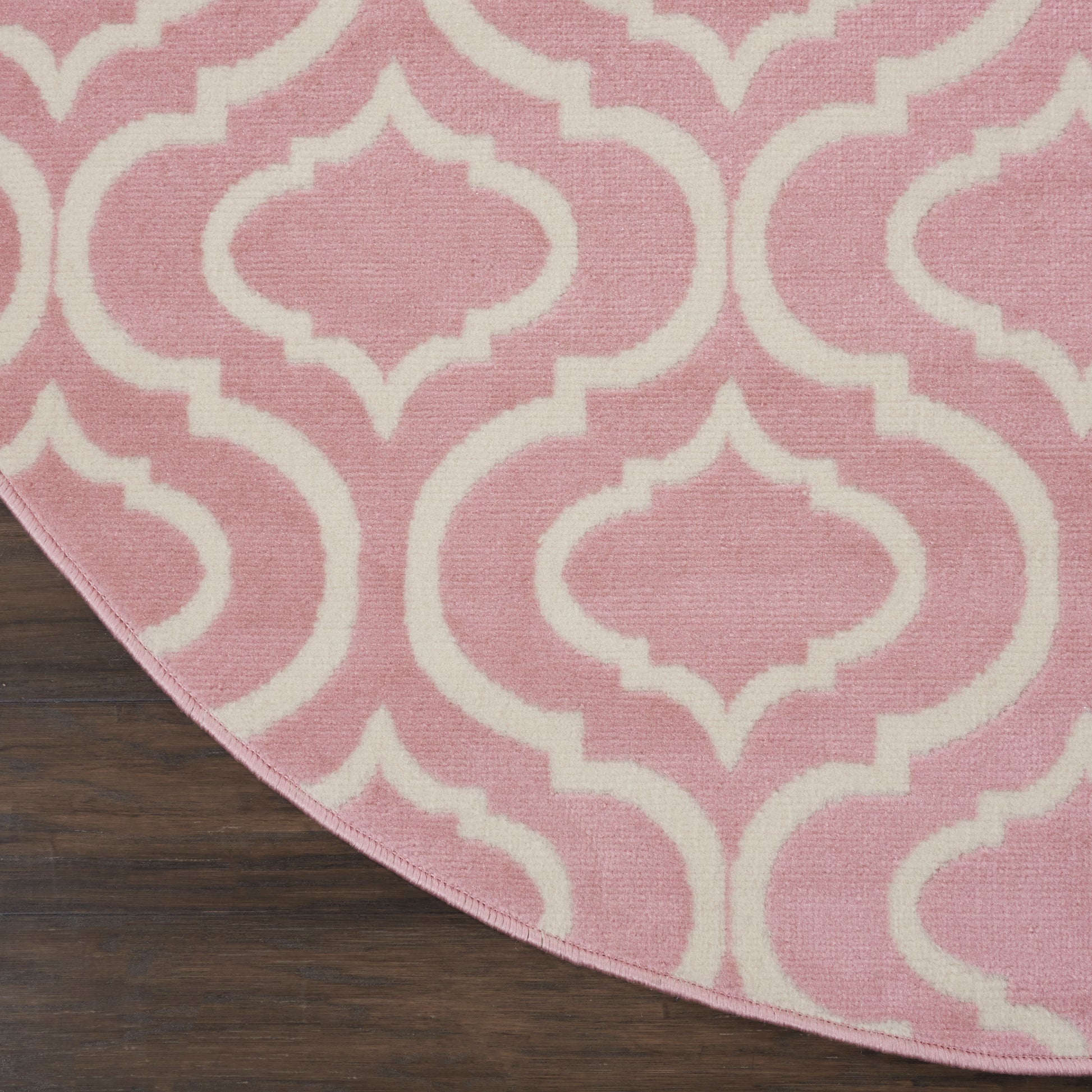 Pink round rug with white trellis pattern; woven texture on dark wood floor.