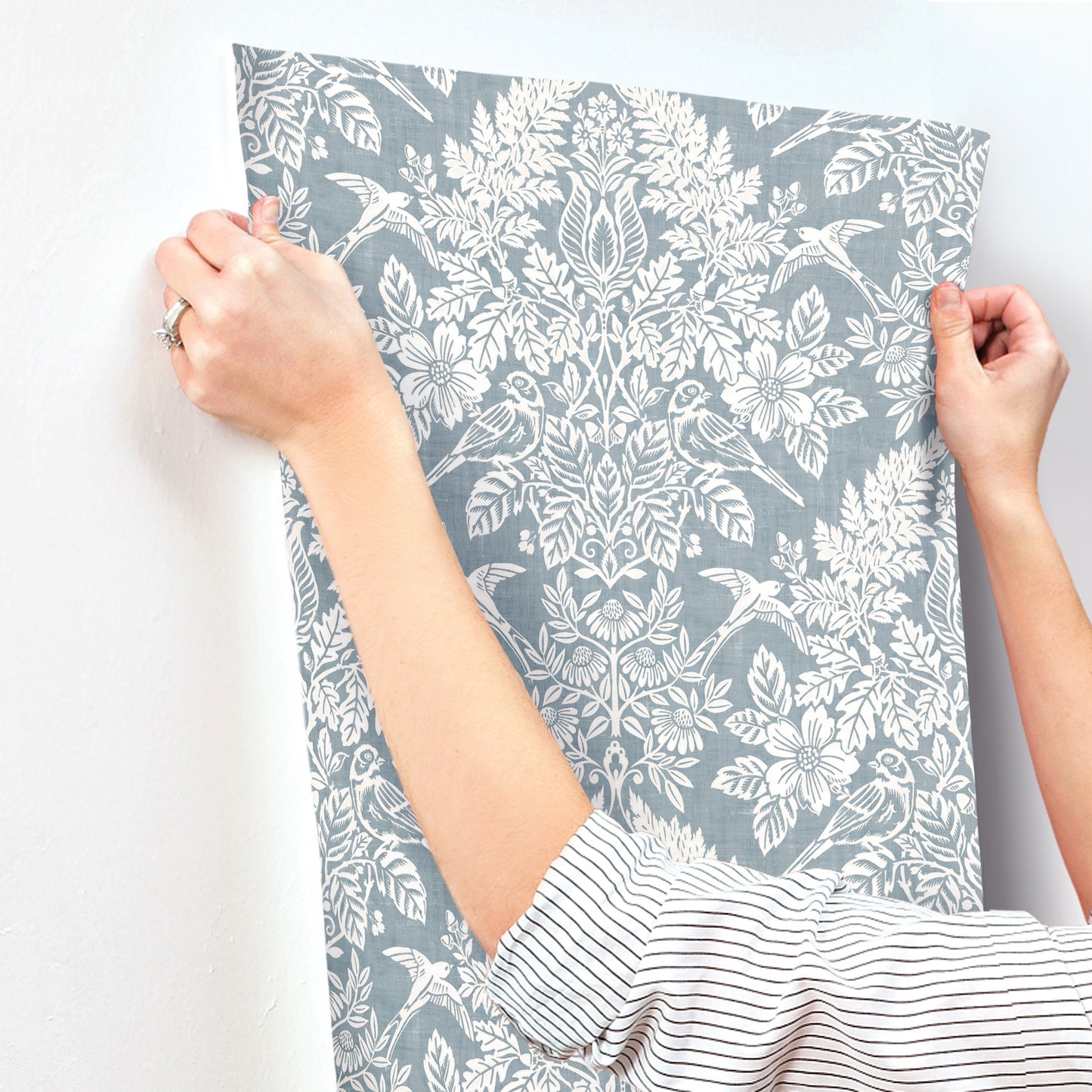 Hands hold a blue-gray wallpaper panel with a white botanical floral pattern; striped shirt sleeve visible.