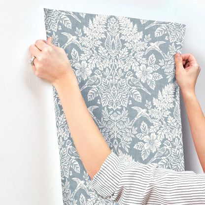 Hands hold a blue-gray wallpaper panel with a white botanical floral pattern; striped shirt sleeve visible.