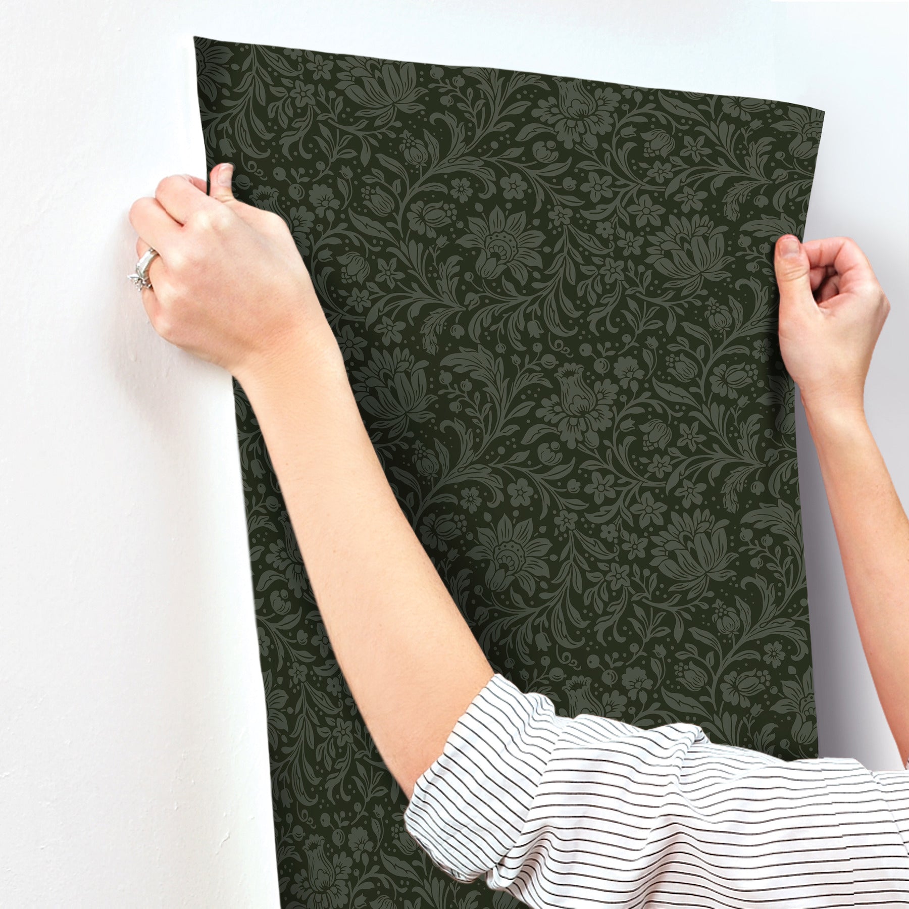 Two hands hold up a dark green damask-patterned wallpaper panel against a white wall.