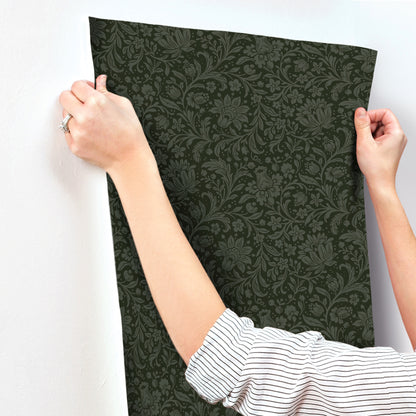 Two hands hold up a dark green damask-patterned wallpaper panel against a white wall.