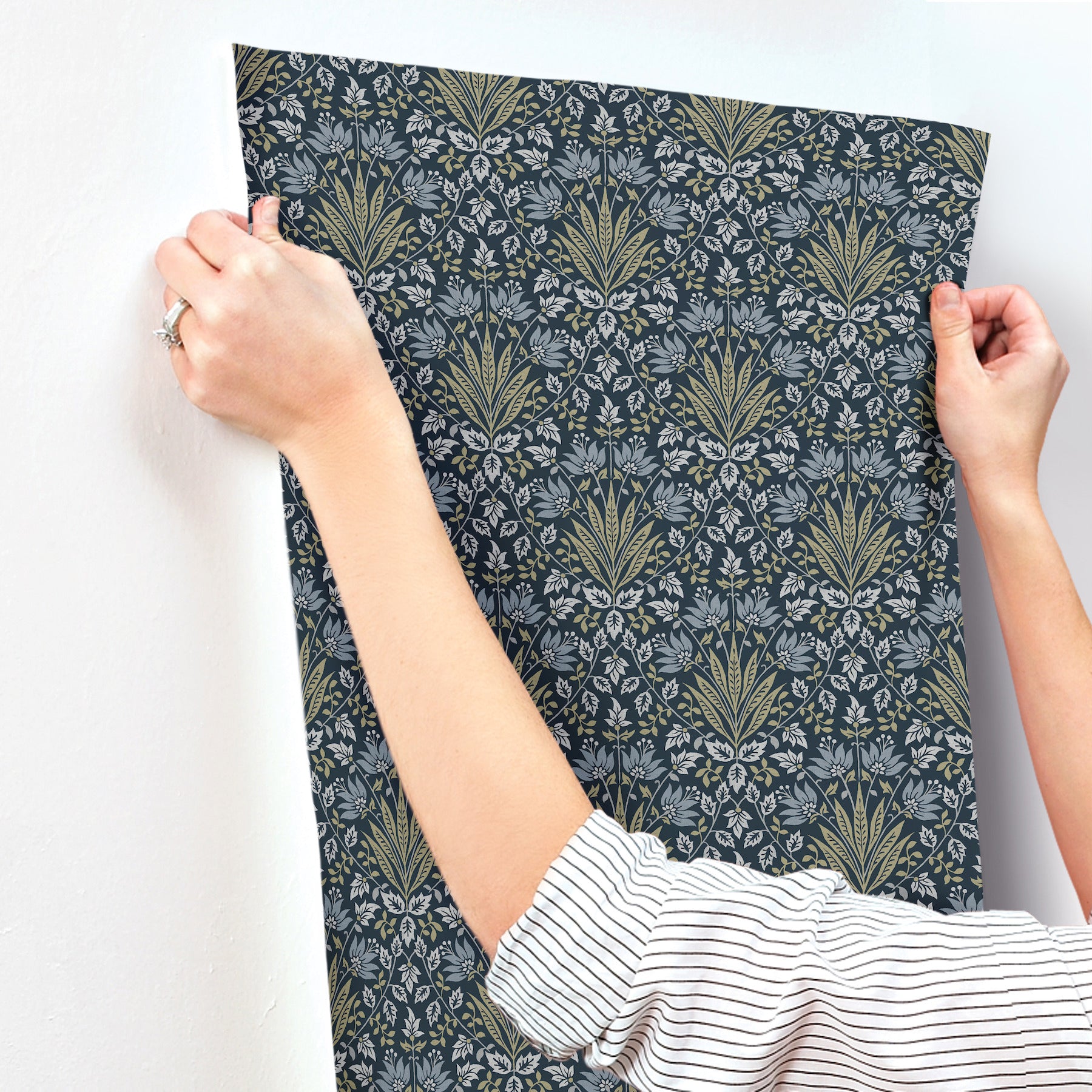 Navy wallpaper swatch with ornate gold and olive damask floral pattern held by hands.