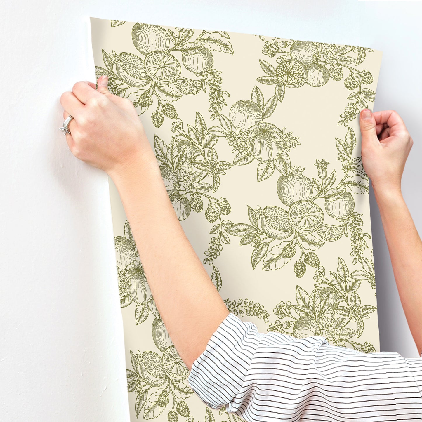 Beige wallpaper panel with a green botanical fruit and leaf pattern, held by hands in striped sleeves.