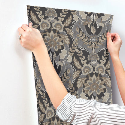 Two hands hold a large rectangular wallpaper sample featuring a charcoal damask floral pattern.