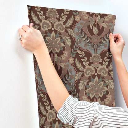 Brown damask floral wallpaper panel with beige and blue-gray motifs held by hands against a white wall.