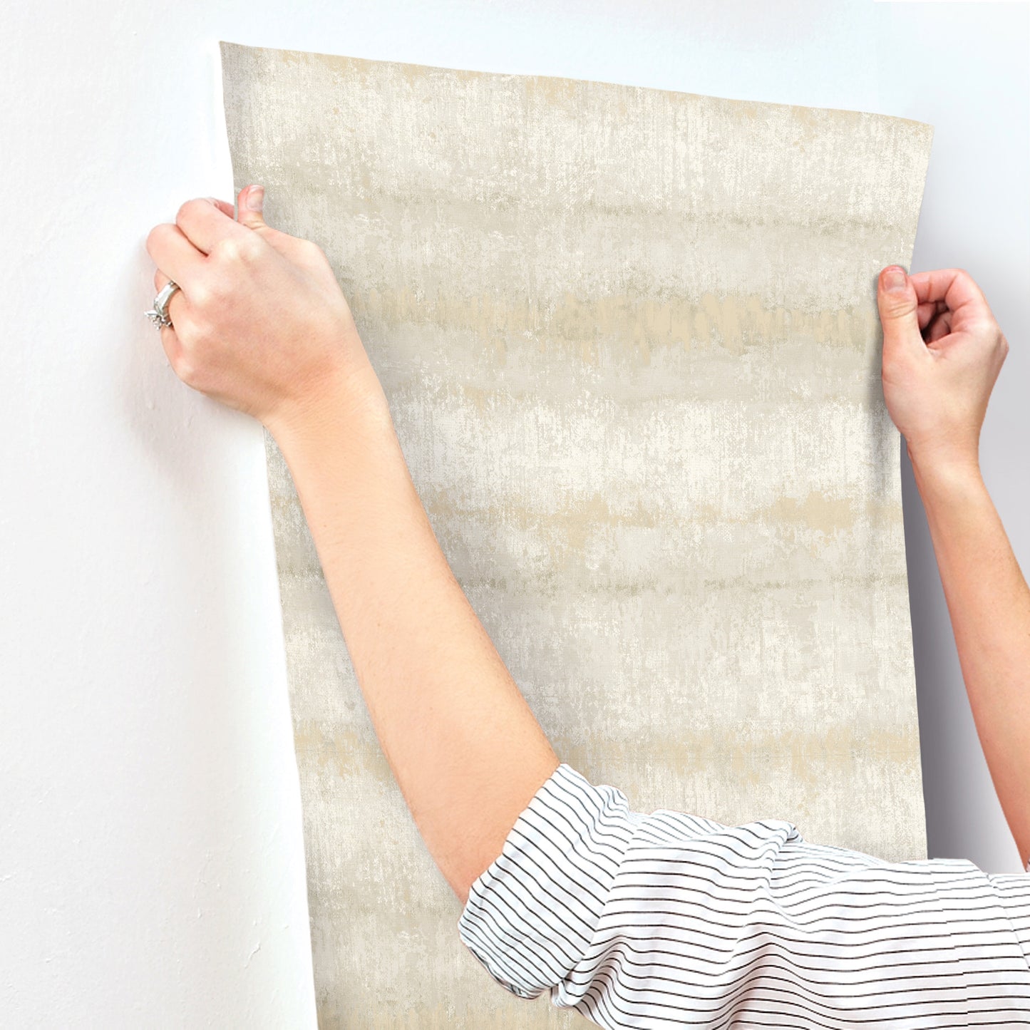 Person hangs a large beige-gray textured striped wallpaper; striped sleeves visible.