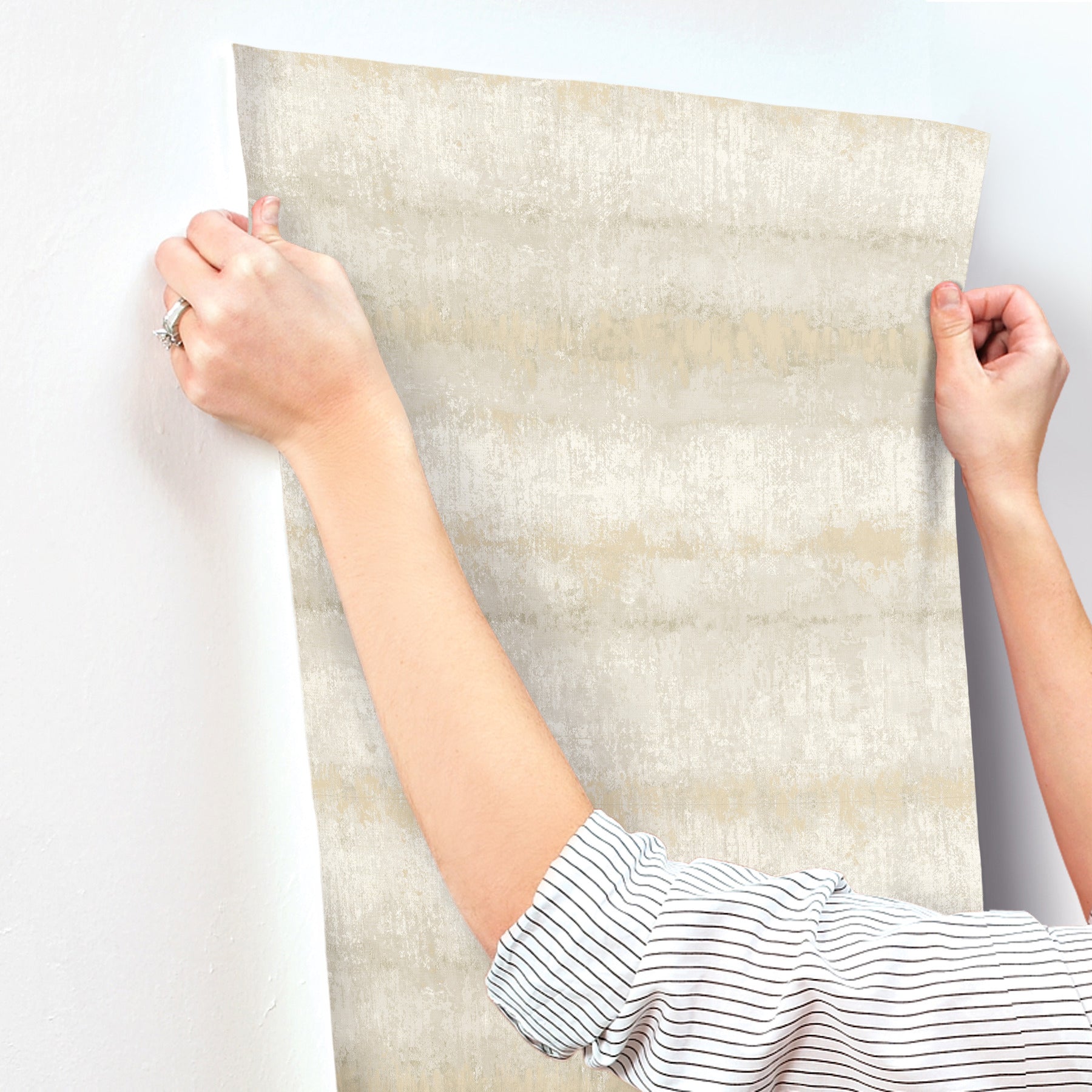 Person hangs a large beige-gray textured striped wallpaper; striped sleeves visible.