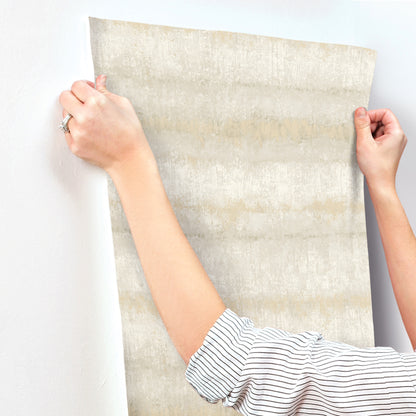 Person hangs a large beige-gray textured striped wallpaper; striped sleeves visible.