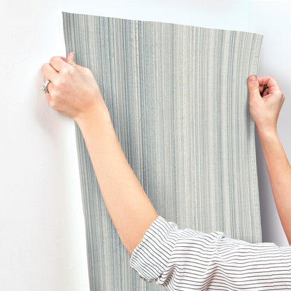 Two hands hold a vertical grey-blue striped wallpaper swatch; striped shirt sleeve visible.