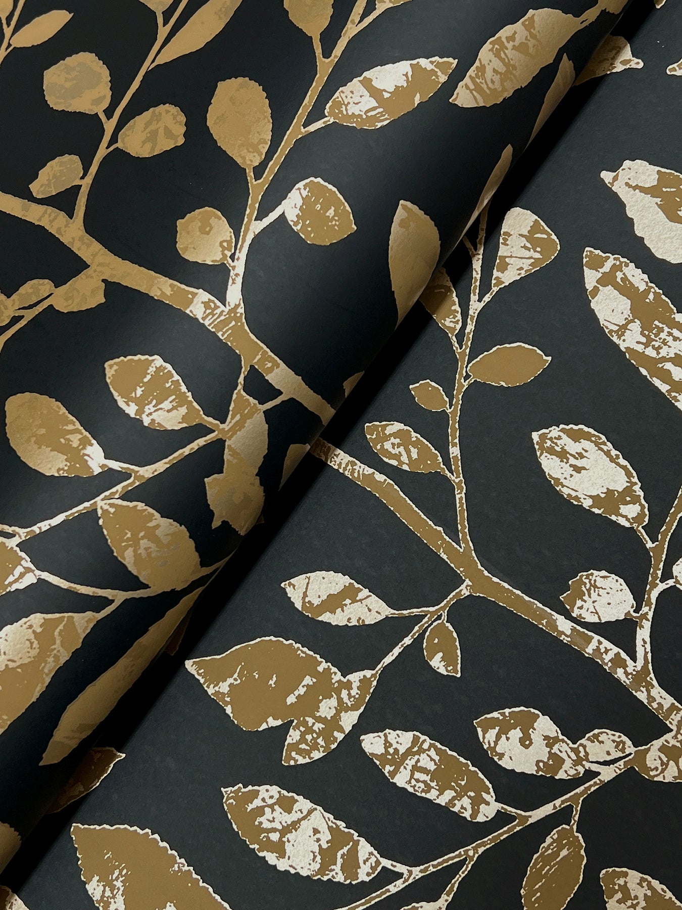 Gold foil botanical branch wallpaper on charcoal background; rolled sheets with metallic leaf pattern.