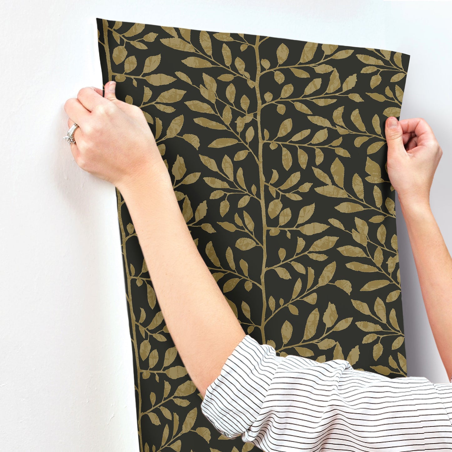 Two hands hold a black wallpaper panel with a gold botanical leaf pattern, elegant nature-inspired wall decor.
